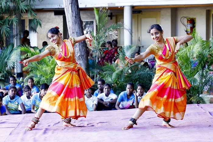 Two young women performing a traditional Indian classical dance on stage in colorful orange and pink costumes, with an audience watching in the background.
