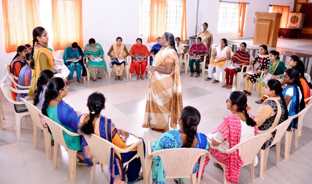 A group of women seated in a circular arrangement in a classroom, participating in an interactive discussion or training session led by a teacher standing in the center.