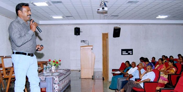 Speaker presenting with a microphone during a seminar at PKR Arts College for Women, while faculty members and students listen in the auditorium.